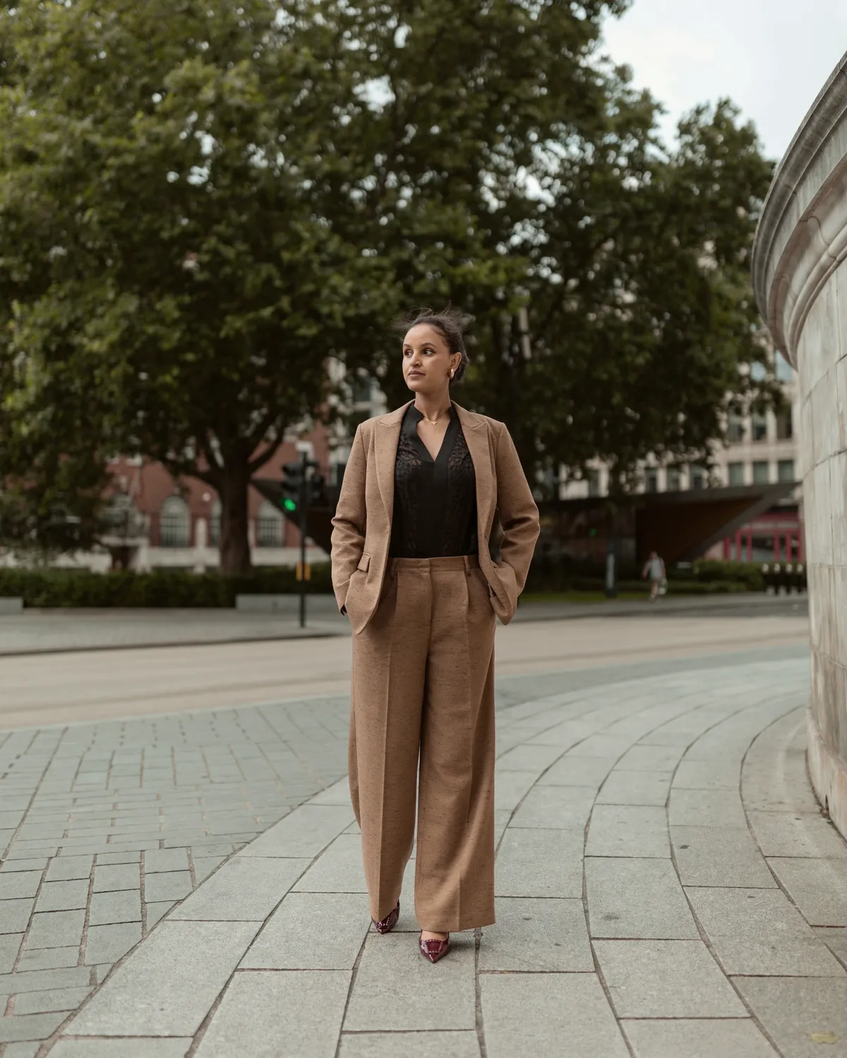 Woman in a tailored camel suit standing on a stone city plaza beneath shade trees